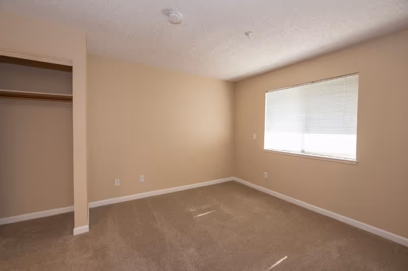 Empty bedroom with beige walls and carpet, a window with closed blinds on the right wall, and an open closet on the left side.