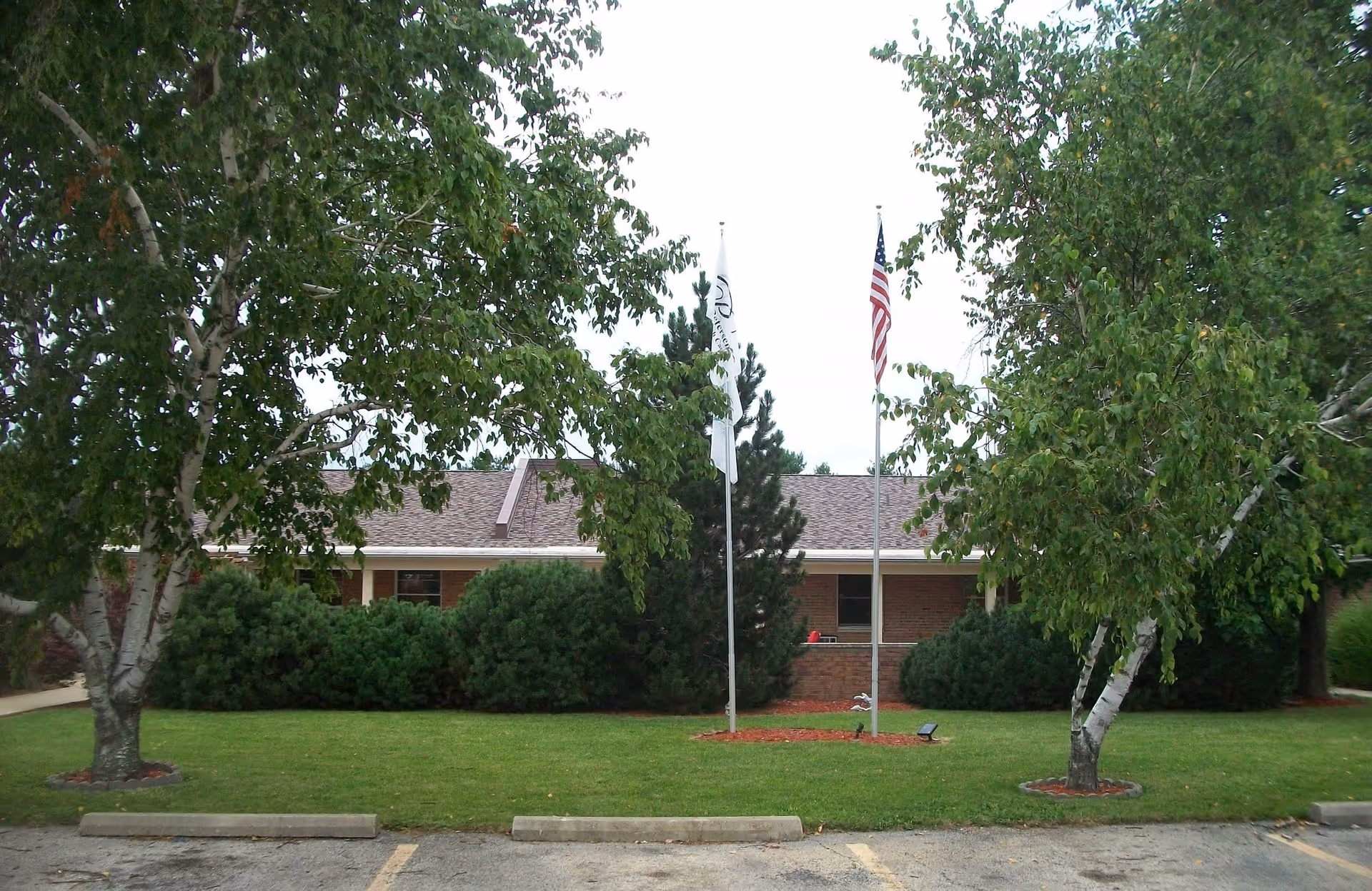 Front view of a single-story brick building partially obscured by trees with a lawn and flagpoles including an American flag.