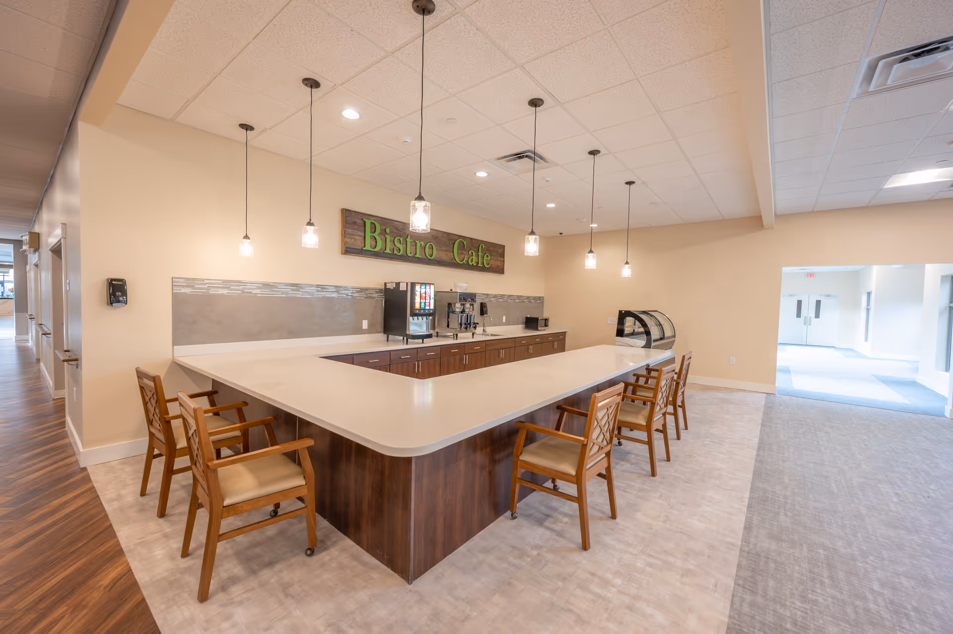 Interior view of a bistro cafe area in a senior living facility with a large U-shaped counter surrounded by wooden chairs with armrests. The counter has a coffee machine, a soda dispenser, and a small display case. Pendant lights hang from the ceiling, and a sign on the wall reads 'Bistro Cafe'. The space is well-lit with a combination of natural and artificial light, and there is a hallway visible to the left and an open area to the right.
