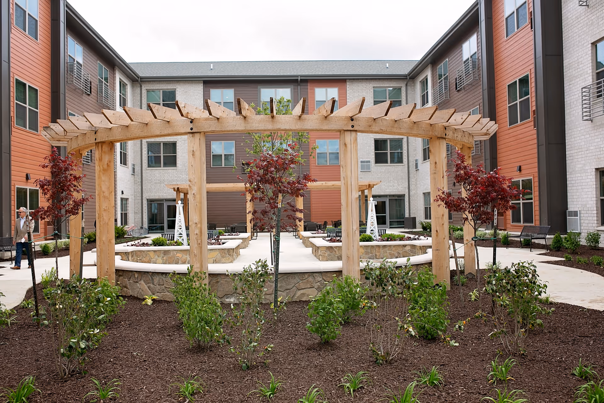 Outdoor courtyard area of a senior living facility with a wooden pergola structure, landscaped garden beds with small trees and shrubs, stone seating walls, and a multi-story building with windows surrounding the courtyard.