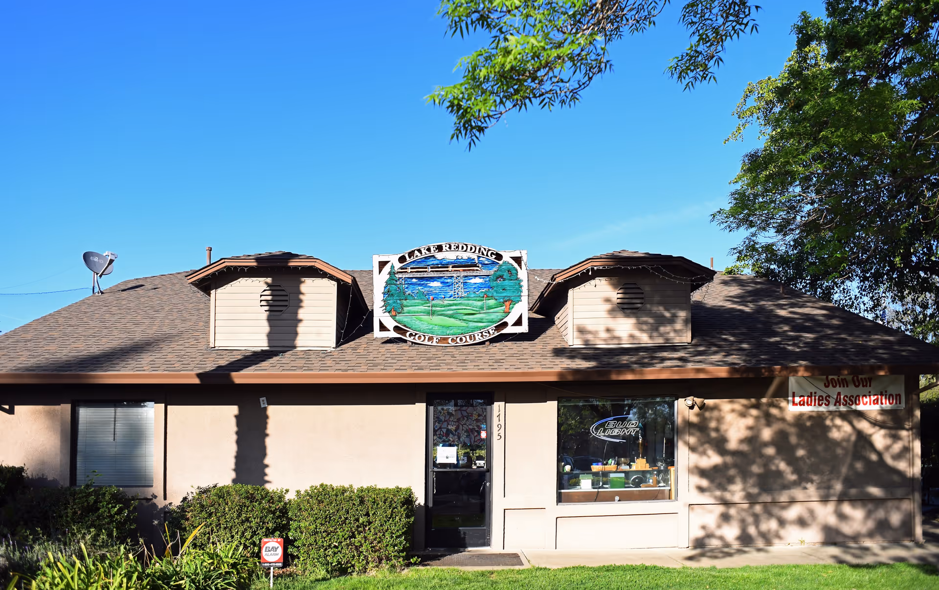 Exterior view of a single-story building with a sign that reads 'Lake Redding Golf Course' mounted above the entrance. The building has a brown roof with two dormer windows and beige walls. There is a glass door and a window displaying a Bud Light sign. A banner on the right side of the building reads 'Join Our Ladies Association'. Green bushes and grass are in front of the building under a clear blue sky.