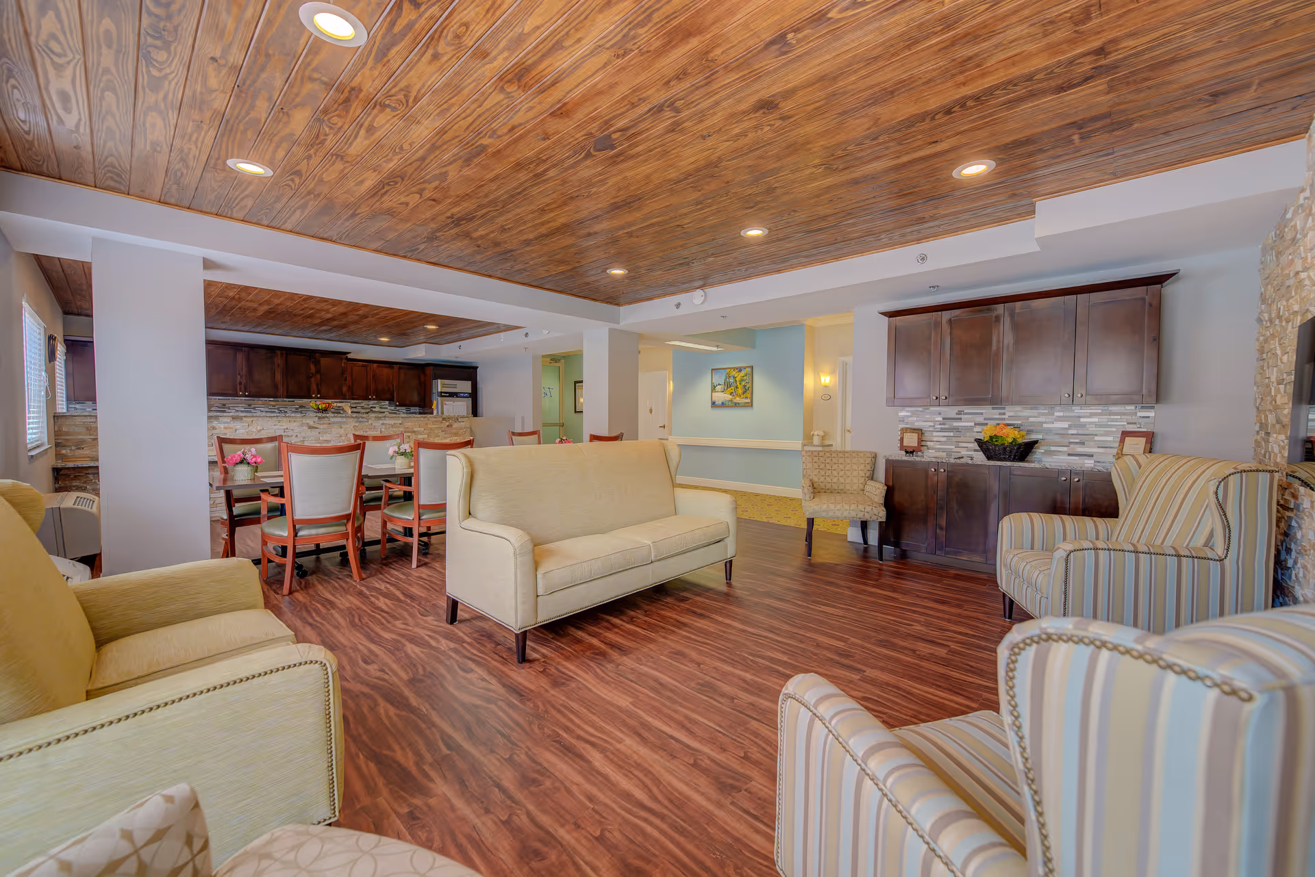 A spacious senior living common area with wooden flooring and ceiling. The room features beige and striped armchairs, a beige sofa, a dining table with chairs, and a kitchen area with dark wooden cabinets and a tiled backsplash. The walls are light-colored, and there is a painting and decorative items on a cabinet.