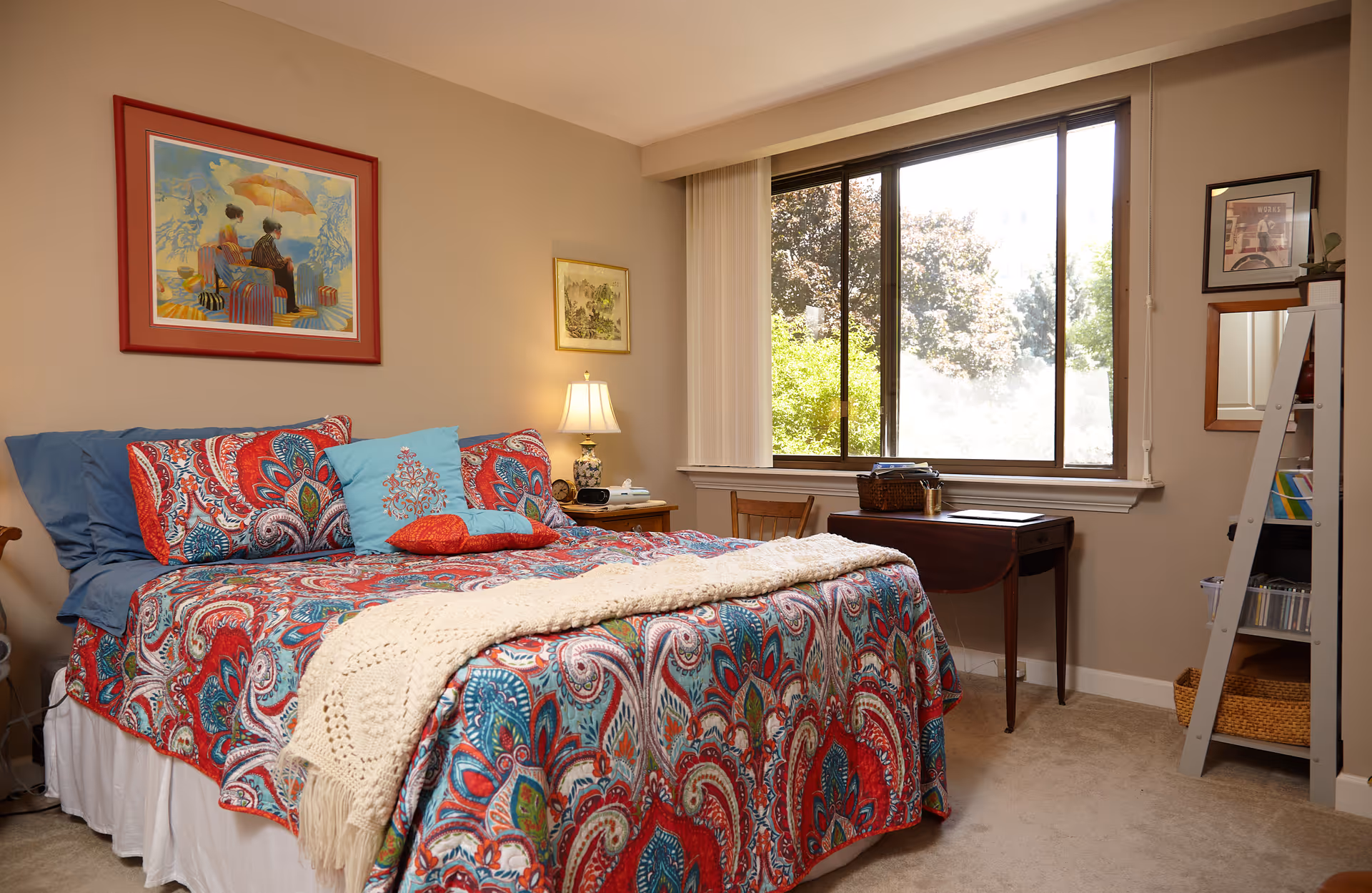 A cozy bedroom with a bed covered in a colorful red, blue, and white patterned comforter and pillows. There is a cream knitted throw blanket at the foot of the bed. A wooden nightstand with a lamp and clock sits next to the bed. A large window with vertical blinds lets in natural light and shows green trees outside. On the right side, there is a small wooden desk with a chair and a white ladder-style shelf with books and baskets. Two framed pictures hang on the beige walls.