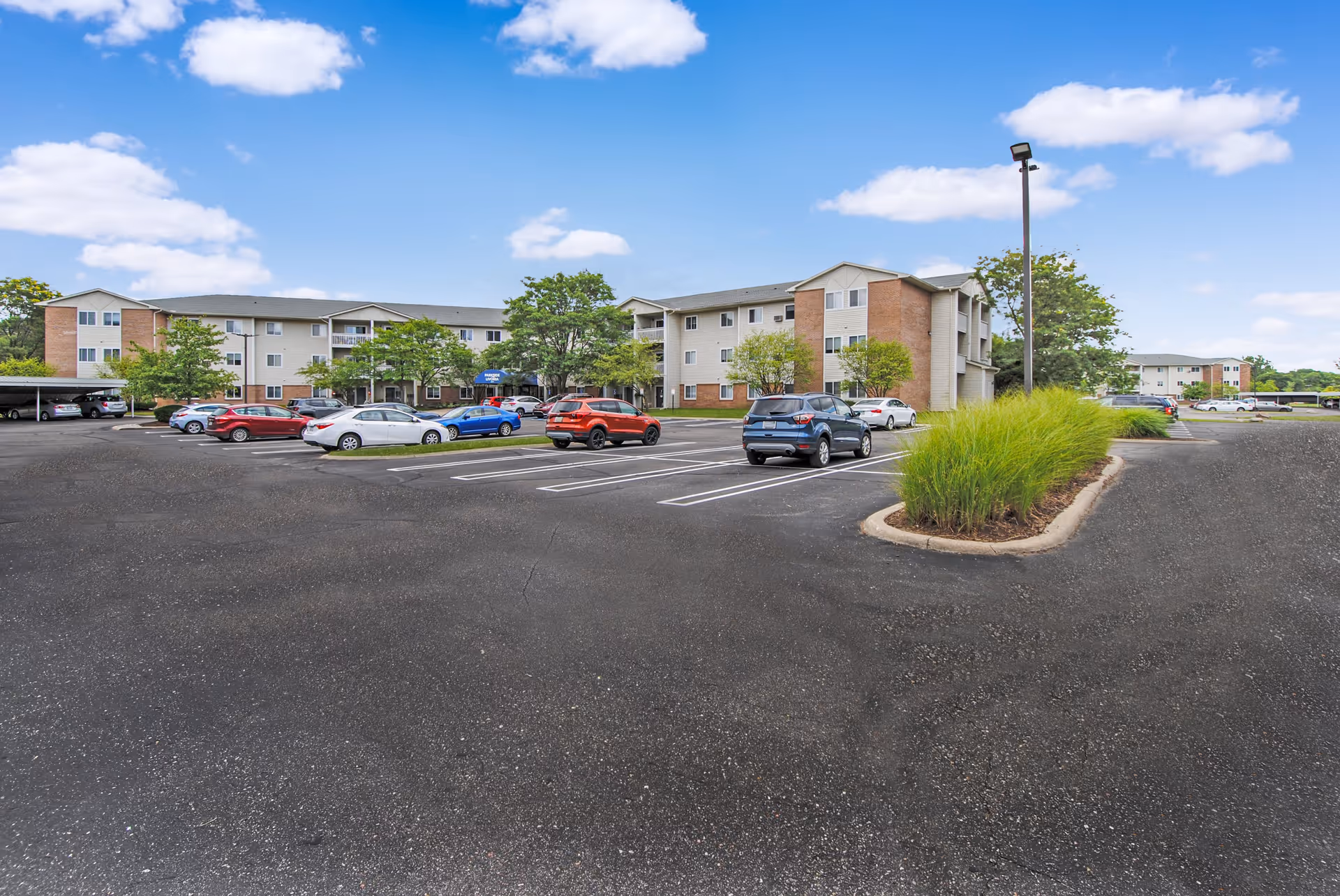 Parking lot with several cars parked in front of a three-story residential building under a blue sky with scattered clouds. The building has a mix of brick and light-colored siding with balconies and surrounded by trees and landscaped areas.