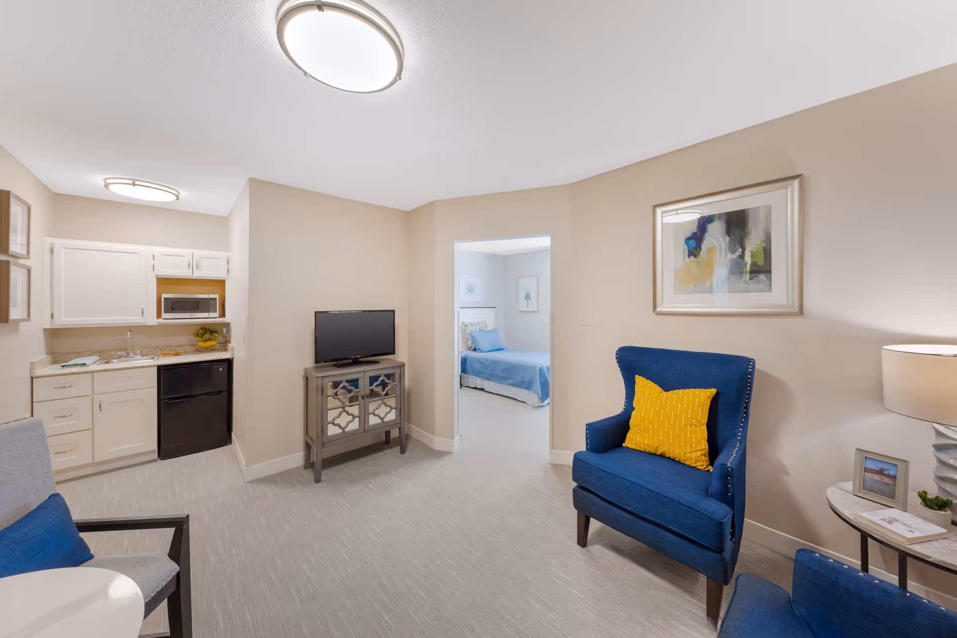 Bright furnished living area with blue armchairs, a small kitchenette, TV on a stand, and a view into a bedroom.