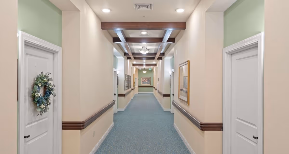 A well-lit, long indoor hallway in a senior living facility with beige walls, light blue carpet, white doors on both sides, wooden handrails, framed artwork on the walls, and decorative wooden beams on the ceiling.