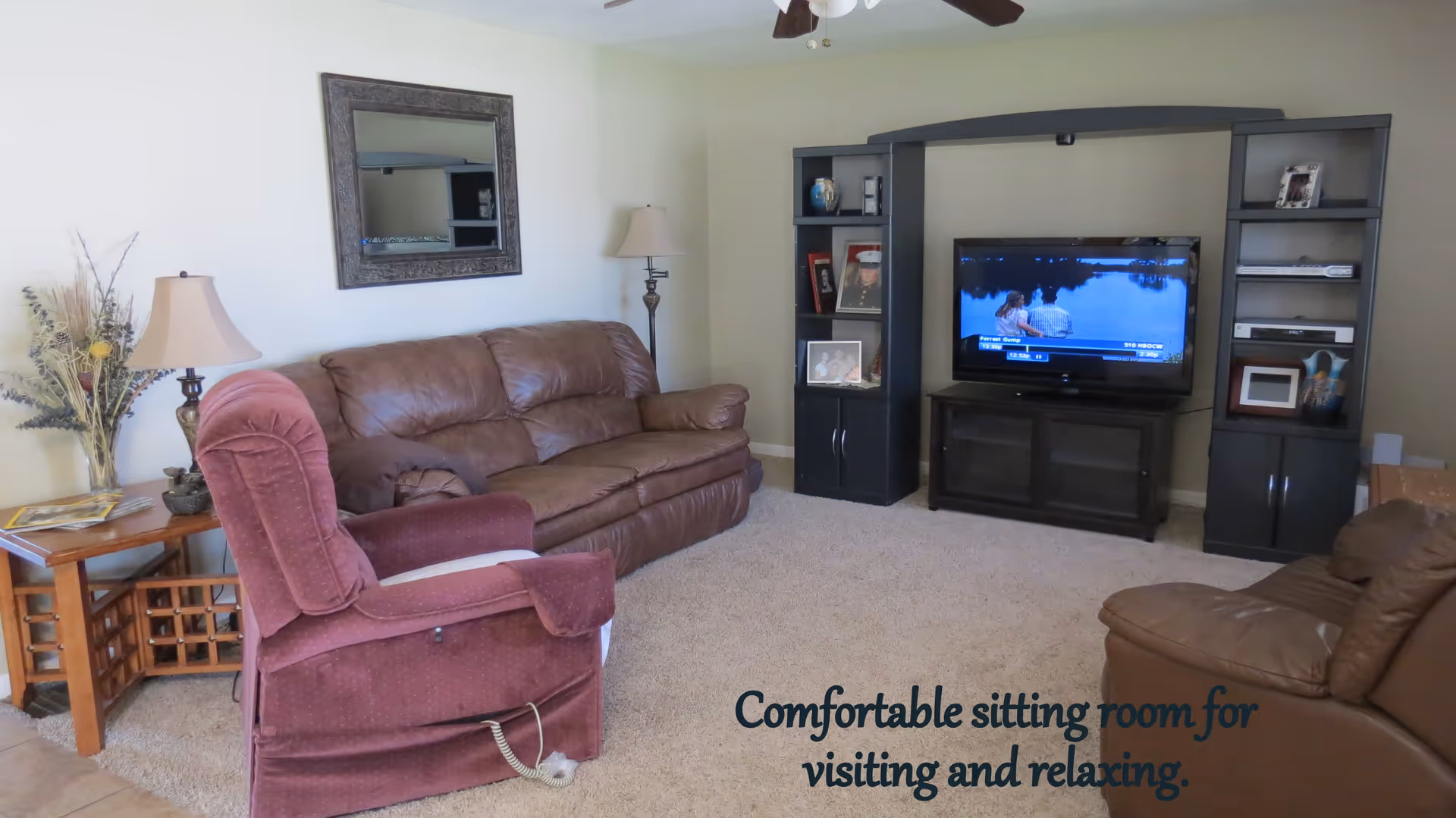 A cozy living room with a brown leather sofa, a maroon recliner chair, a wooden side table with a lamp and decorative dried plants, a large mirror on the wall, and a black entertainment center with a flat-screen TV showing a scene from the movie Forrest Gump. The room has beige carpet and light-colored walls.