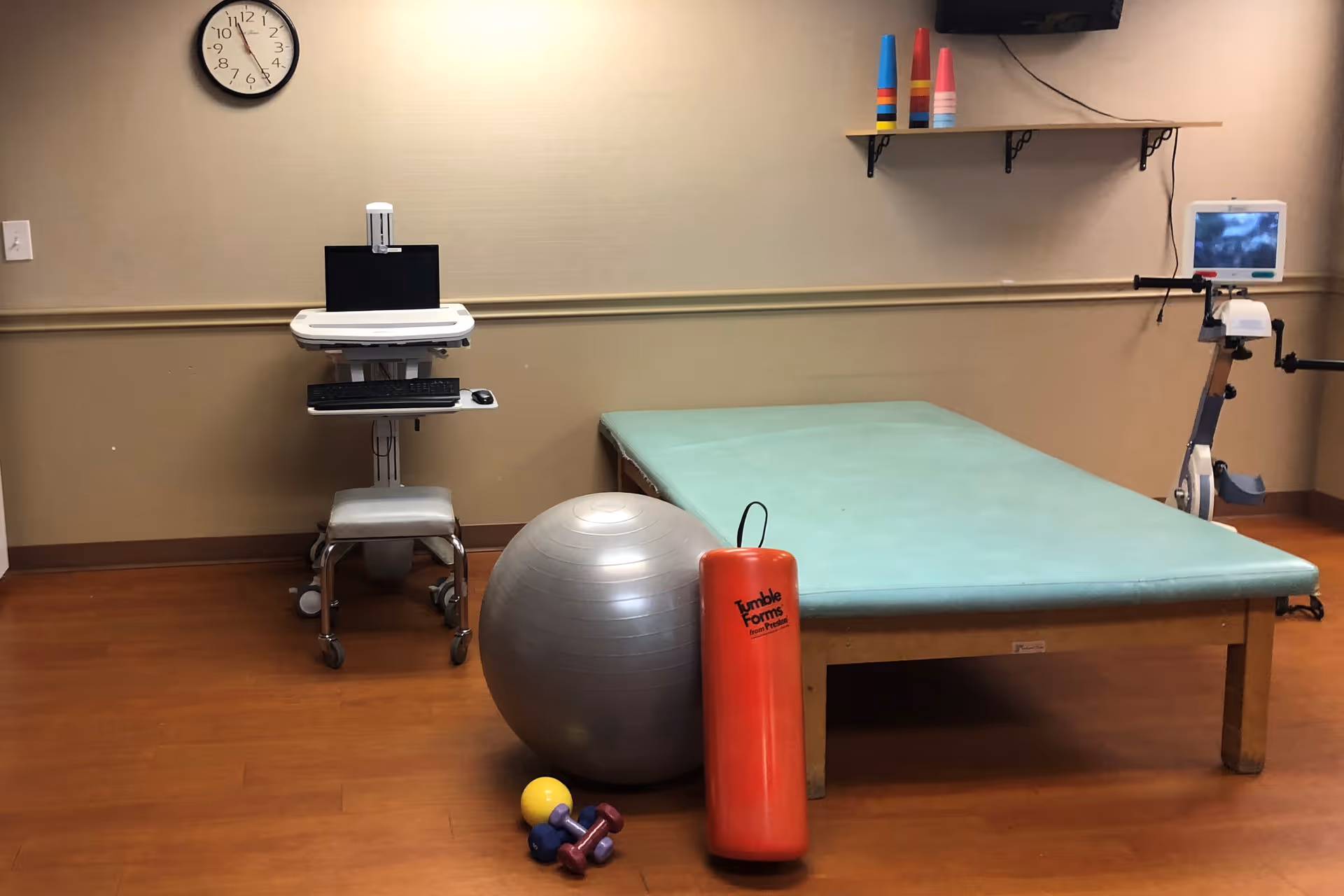 Physical therapy room with a padded treatment table, exercise ball, small dumbbells, a red foam roller, a computer workstation, and an exercise bike.