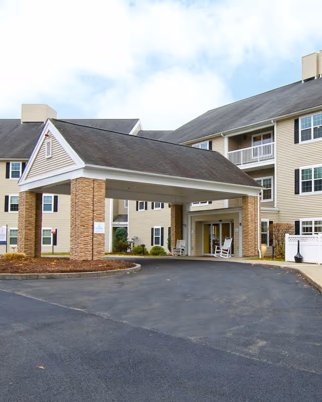 Entrance of a senior living facility with a covered drop-off area supported by stone pillars. The building has beige siding, multiple windows with black shutters, and a few balconies. There are two white rocking chairs near the glass entrance doors. The sky is partly cloudy.