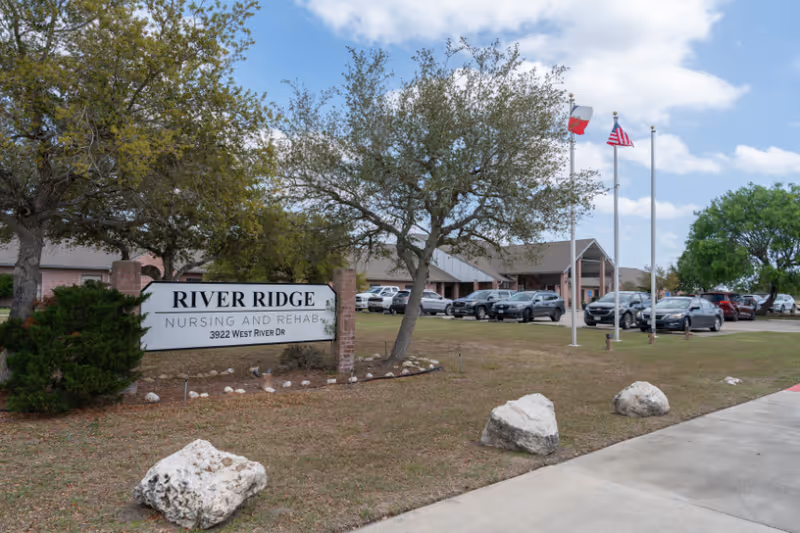 Exterior view of River Ridge Nursing and Rehabilitation facility with a sign displaying the name and address. The building is surrounded by trees, a parking lot with several cars, and three flagpoles with flags against a partly cloudy sky.
