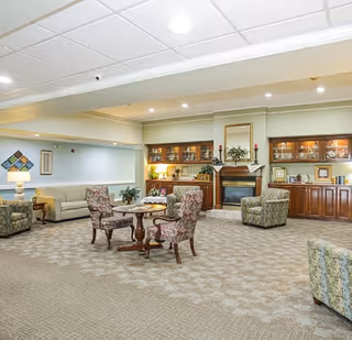 A spacious and well-lit living room area with patterned carpet, several upholstered armchairs and sofas arranged around a central wooden table. The room features a fireplace with a mirror above it, built-in wooden cabinets with glass doors displaying decorative items, and soft lighting from lamps and ceiling lights.