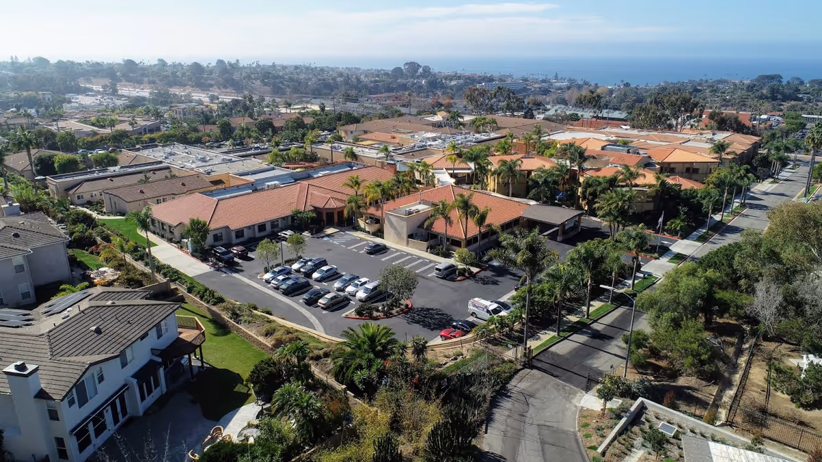 Aerial view of a retirement community campus with red-tiled buildings, a parking lot, palm trees and the ocean visible on the horizon.