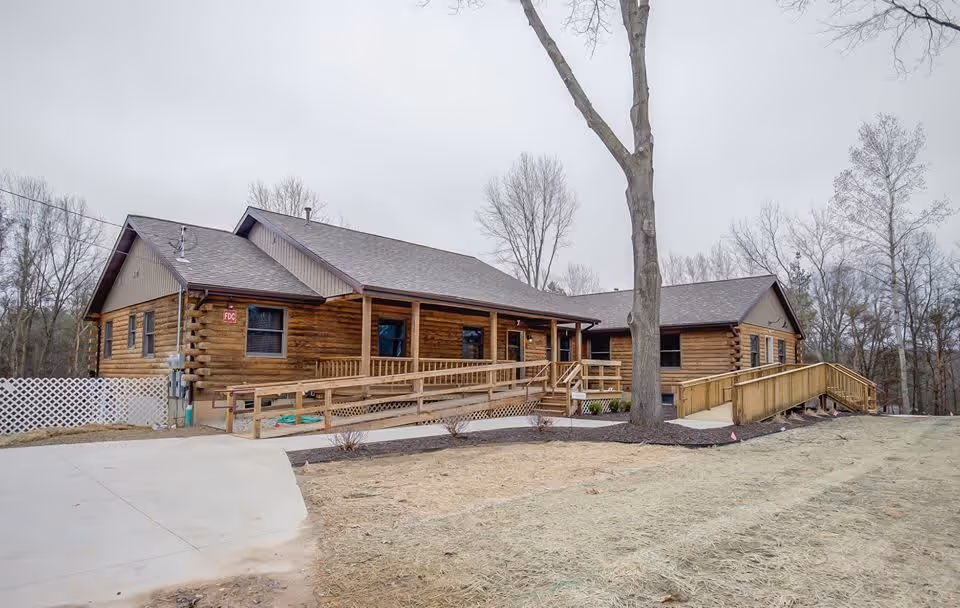 Exterior view of a single-story wooden building with a sloped roof, featuring two wheelchair ramps leading to the entrances. The building is surrounded by bare trees and a patch of grass with some mulch beds. The sky is overcast.