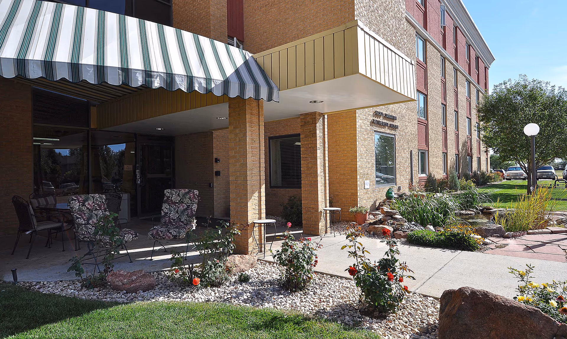 Exterior view of Good Samaritan Society - Fort Collins Village showing the entrance area with a striped awning, patio chairs with floral cushions, a small garden with flowers and rocks, and a pond with plants. The building is a multi-story brick structure with windows and a well-maintained lawn and trees nearby.