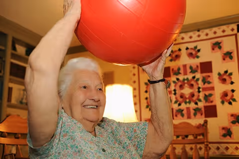 An elderly woman smiling and holding a large red exercise ball above her head in a cozy room with wooden furniture, a lamp, and a floral quilt hanging on the wall.