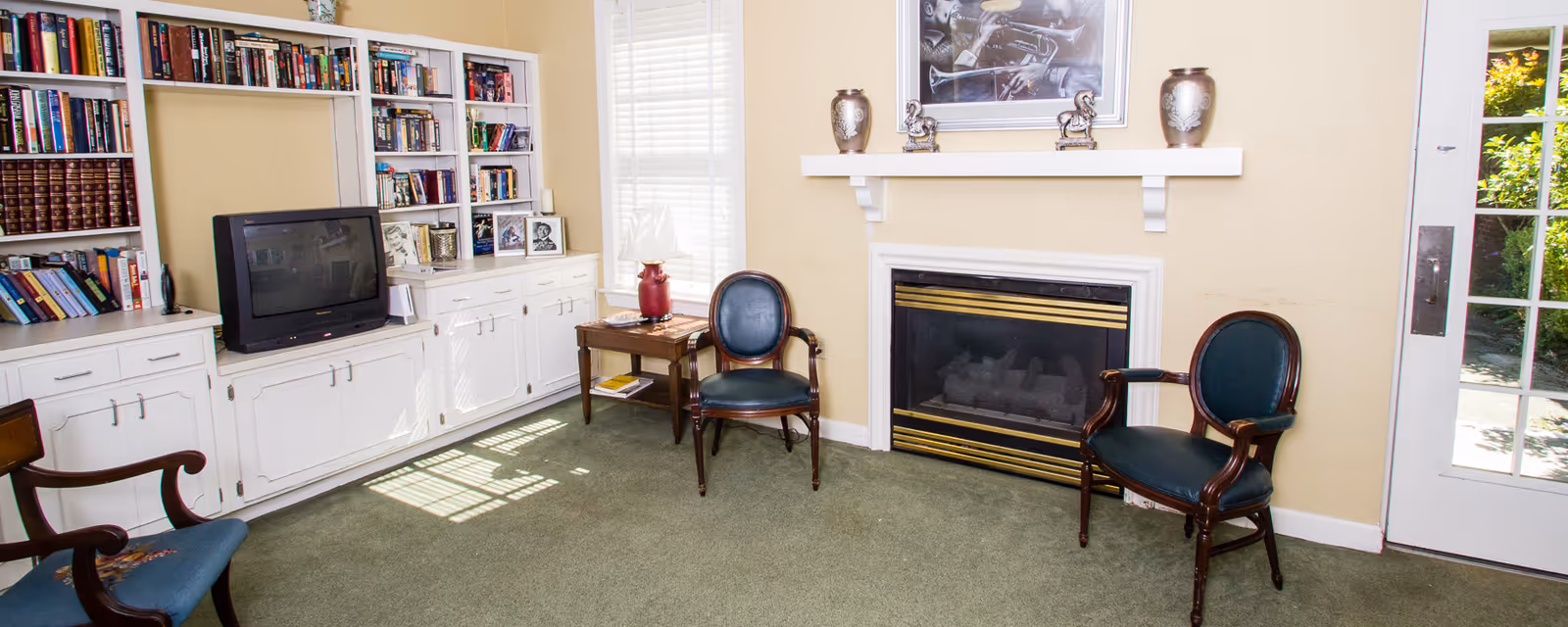 Sunlit common room with a fireplace, built-in bookshelves and cabinets, a TV, and several armchairs.