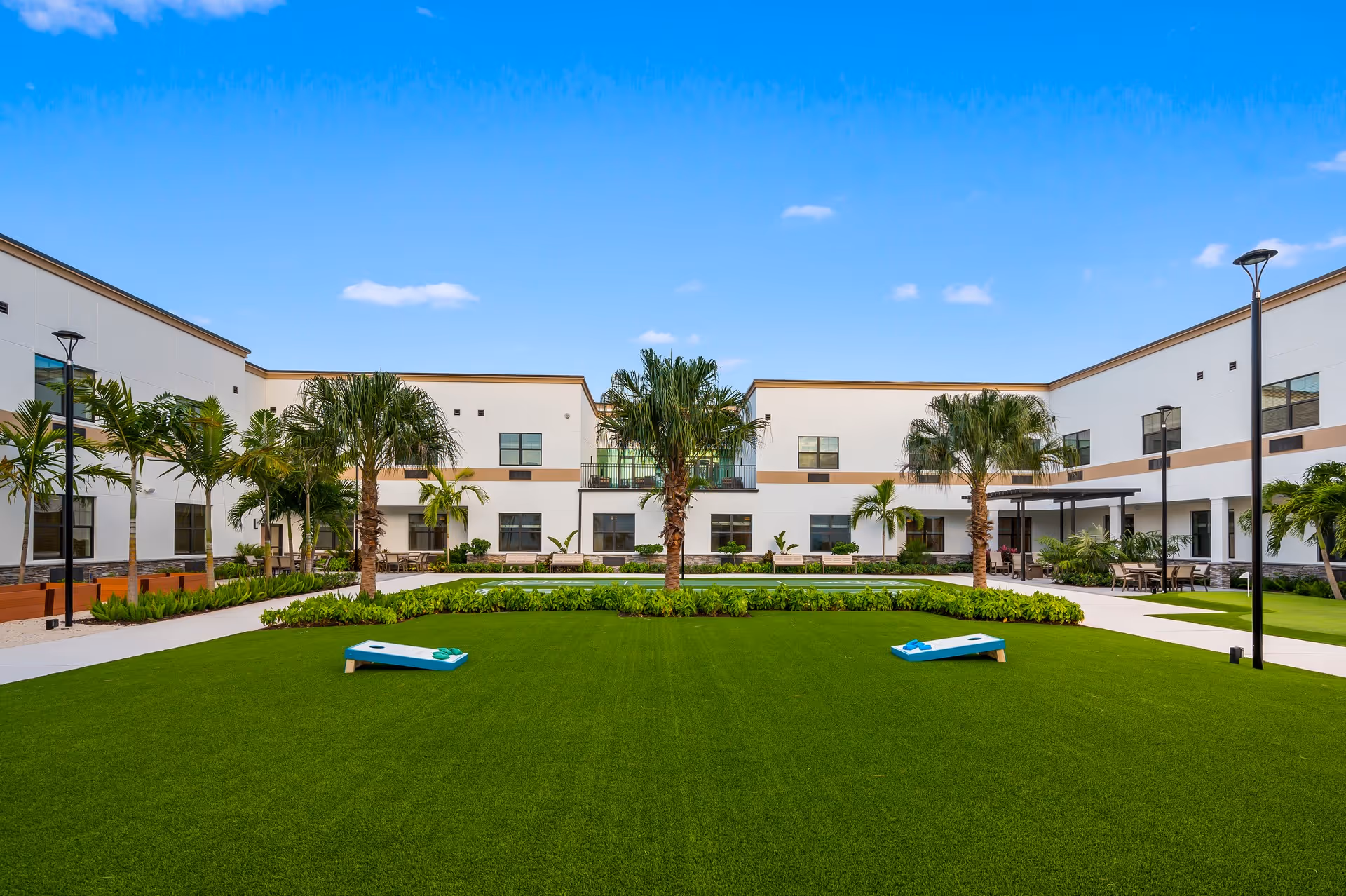 Outdoor courtyard area of Bella Mar Royal Palm Beach facility with green grass, palm trees, and two cornhole boards set up for a game. The courtyard is surrounded by a two-story white building with windows and outdoor seating areas under a clear blue sky.