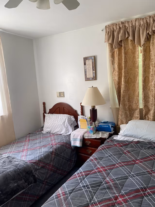 A small bedroom with two twin beds featuring plaid bedding, a nightstand with a lamp and tissues, and a window with patterned curtains.