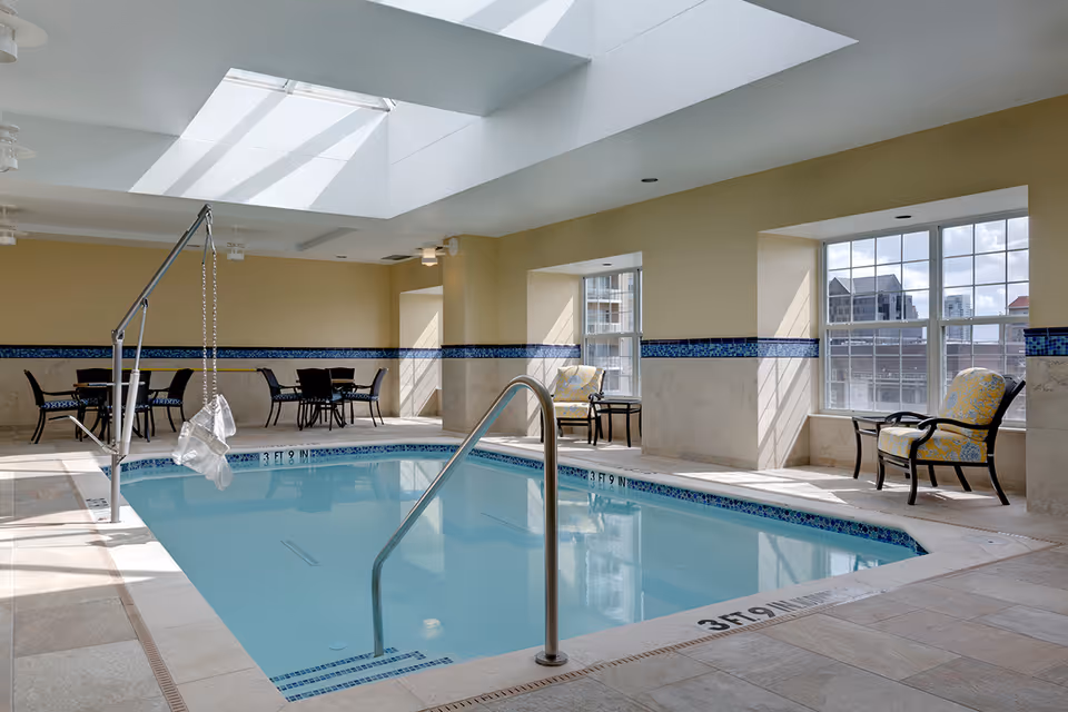 Indoor swimming pool area with a metal handrail and a pool lift for accessibility. The room has large windows letting in natural light, two cushioned chairs with a small table between them, and a few tables with chairs in the background. The walls are painted light yellow with a decorative blue tile border.