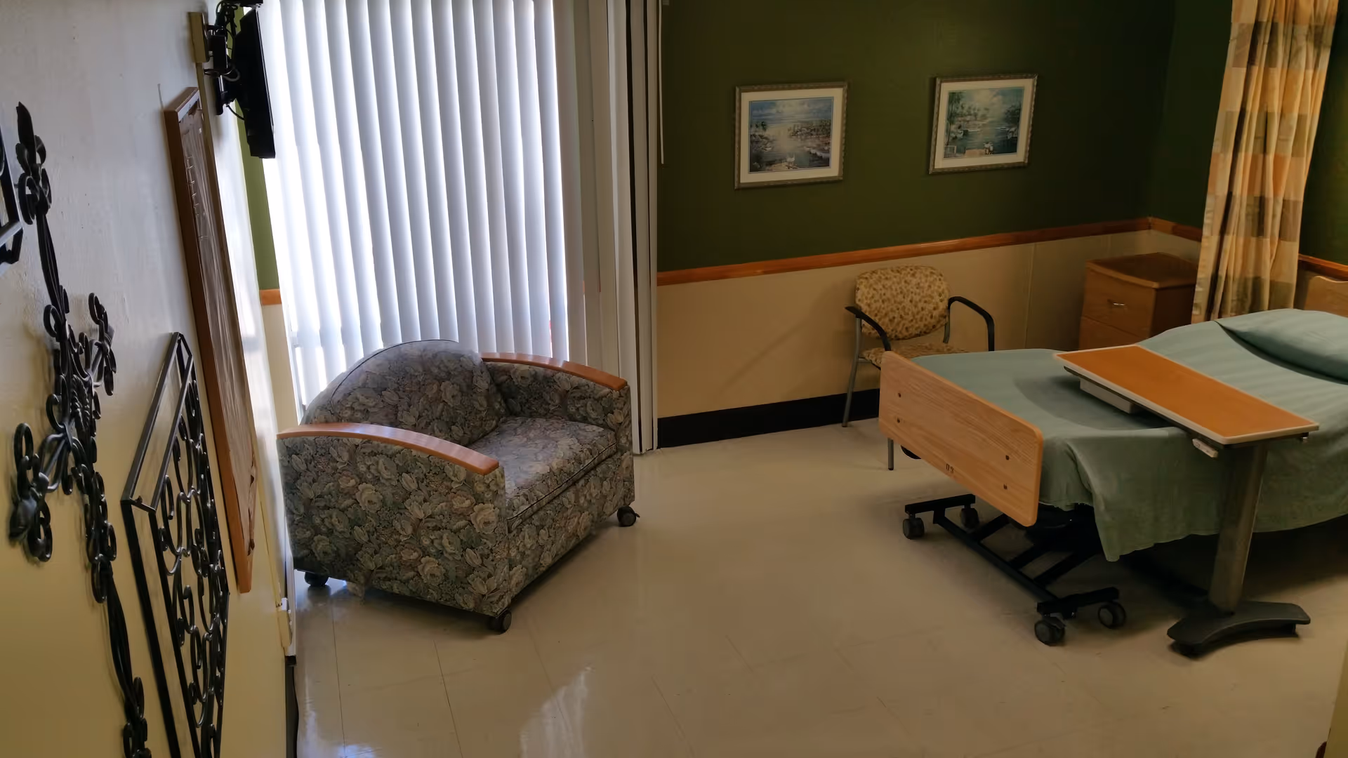 Patient room with a hospital bed and overbed table, a floral armchair, a small side chair and dresser, and vertical blinds over a window.