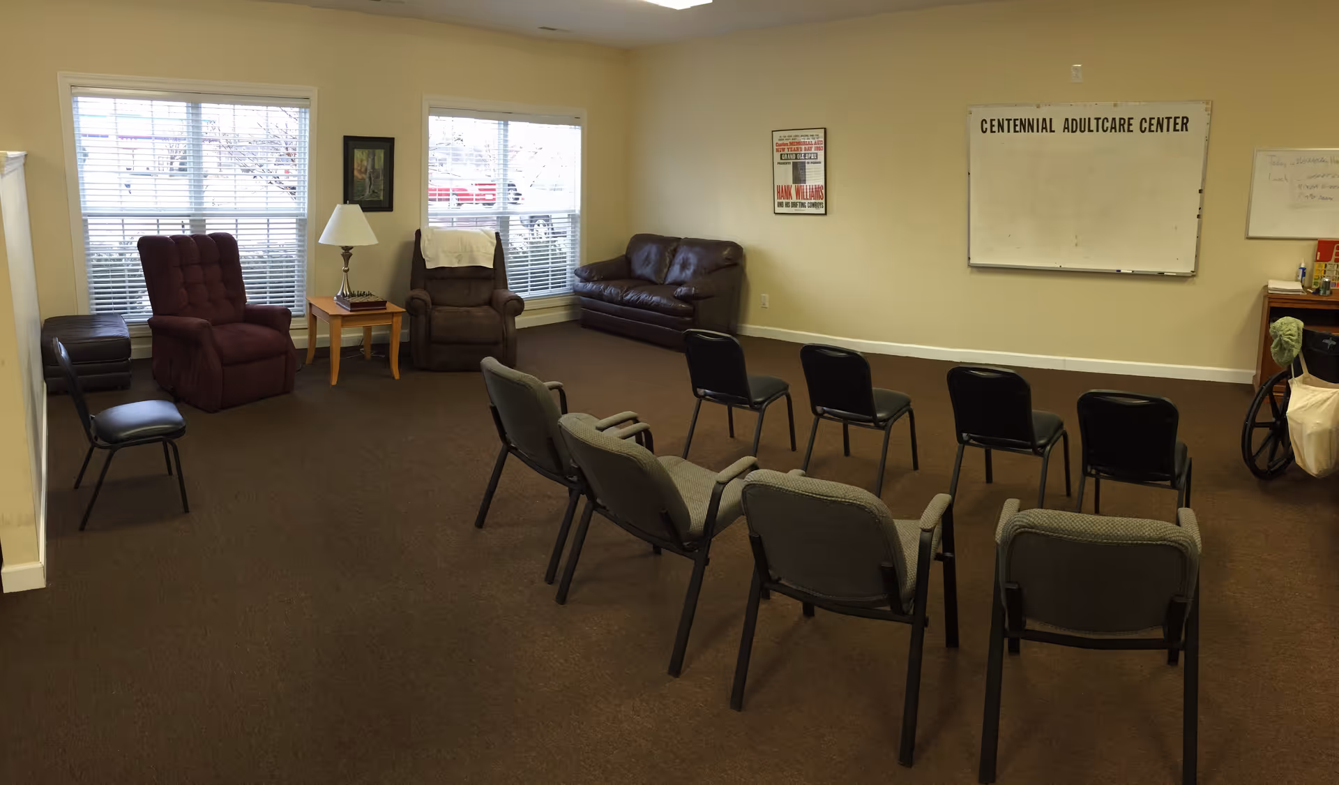 A room with multiple chairs arranged in rows facing a whiteboard labeled 'Centennial Adultcare Center'. The room has two large windows with blinds, a few armchairs, a side table with a lamp, and a small cabinet with a wheelchair nearby. The walls are light-colored and the floor is carpeted.