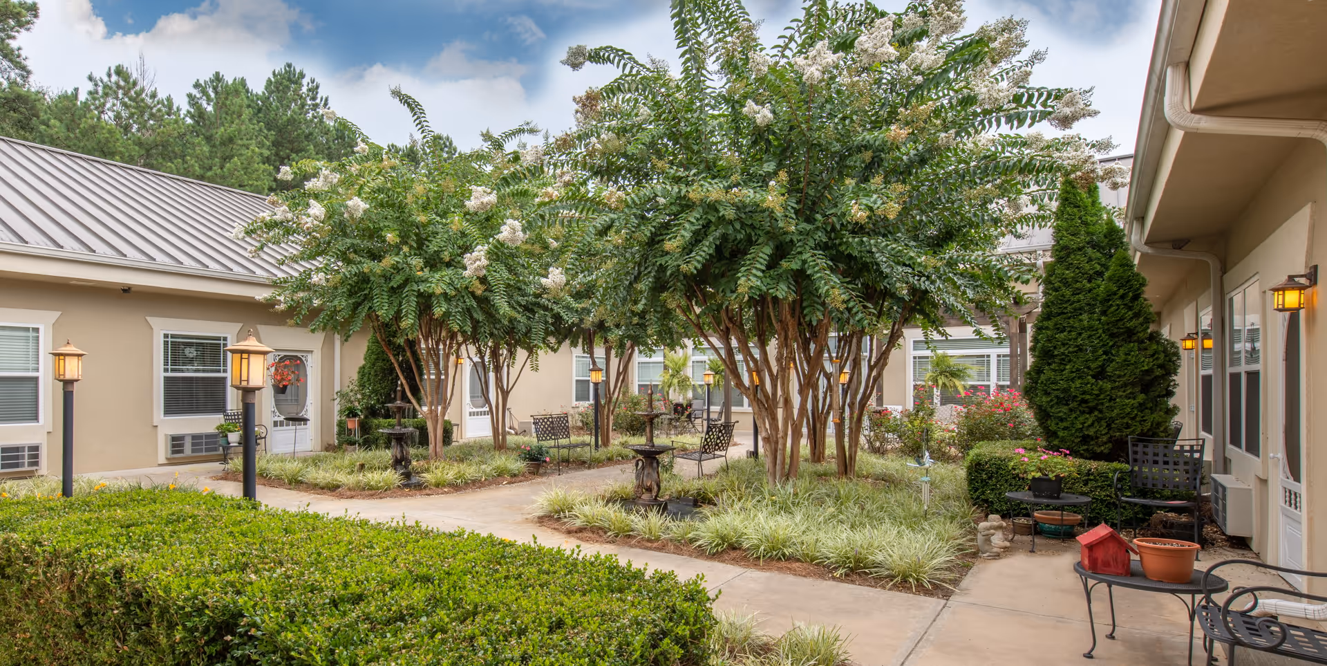 Outdoor courtyard area at Benton House of Newnan featuring a paved walkway surrounded by green shrubs, trees with white flowers, benches, and outdoor seating. The courtyard is enclosed by beige buildings with windows and doors, and there are lamp posts providing lighting.