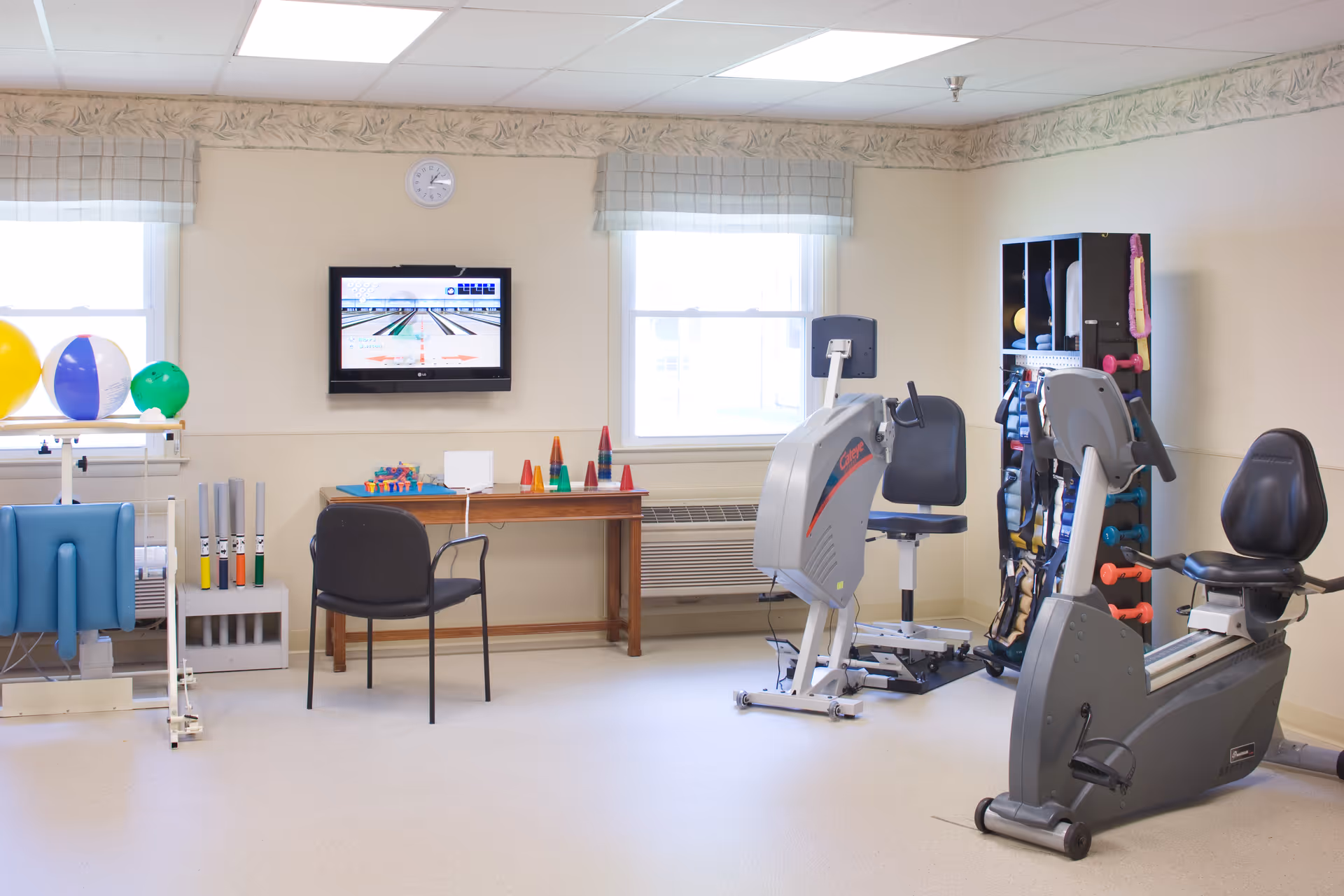 A bright exercise room with two stationary exercise bikes, a rack with various exercise equipment including dumbbells and resistance bands, a table with colorful cones and toys, a chair, and a wall-mounted TV displaying a bowling game. Two windows with light-colored curtains let in natural light.