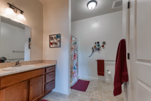 A bathroom with a wooden vanity featuring a sink and a large mirror above it. The floor is tiled, and there is a shower with a floral curtain. Red towels hang on the wall and door, and decorative wall art is displayed above the towel rack.
