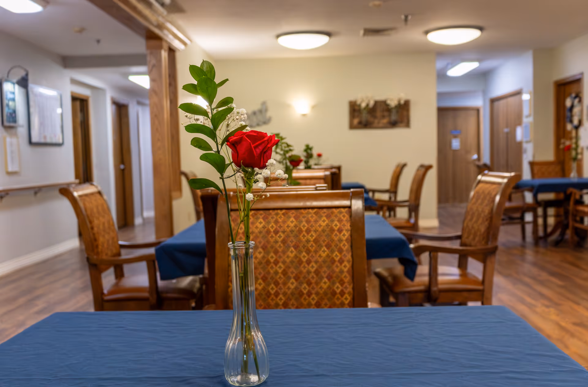 Interior view of a dining area in a senior living facility with wooden chairs and tables covered with blue tablecloths. A clear glass vase with a single red rose and greenery is placed on the closest table. The room has wooden flooring, beige walls, and several doors and wall decorations in the background.