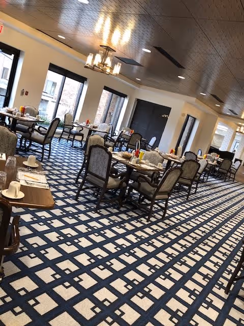 A spacious dining room with multiple tables and upholstered chairs arranged neatly. The room features large windows allowing natural light to enter, a patterned carpet with a geometric design, and modern ceiling lights including chandeliers. Tables are set with cups, plates, and condiments, ready for a meal.