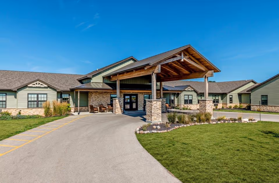 Exterior view of The Lodge at Manito senior living facility showing a single-story building with green siding, stone accents, and a covered entrance with wooden beams. The driveway curves in front of the entrance, and there is a well-maintained lawn with landscaping under a clear blue sky.