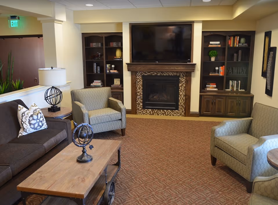 A cozy living room area in a senior living facility featuring a brown sofa with patterned pillows, two light green armchairs, a wooden coffee table with decorative items, a fireplace with a mosaic tile surround, built-in wooden bookshelves filled with books and decor, and a large flat-screen TV mounted above the fireplace.