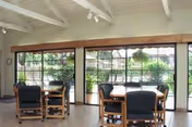 Bright communal dining area with tables and chairs in front of large windows overlooking a garden.