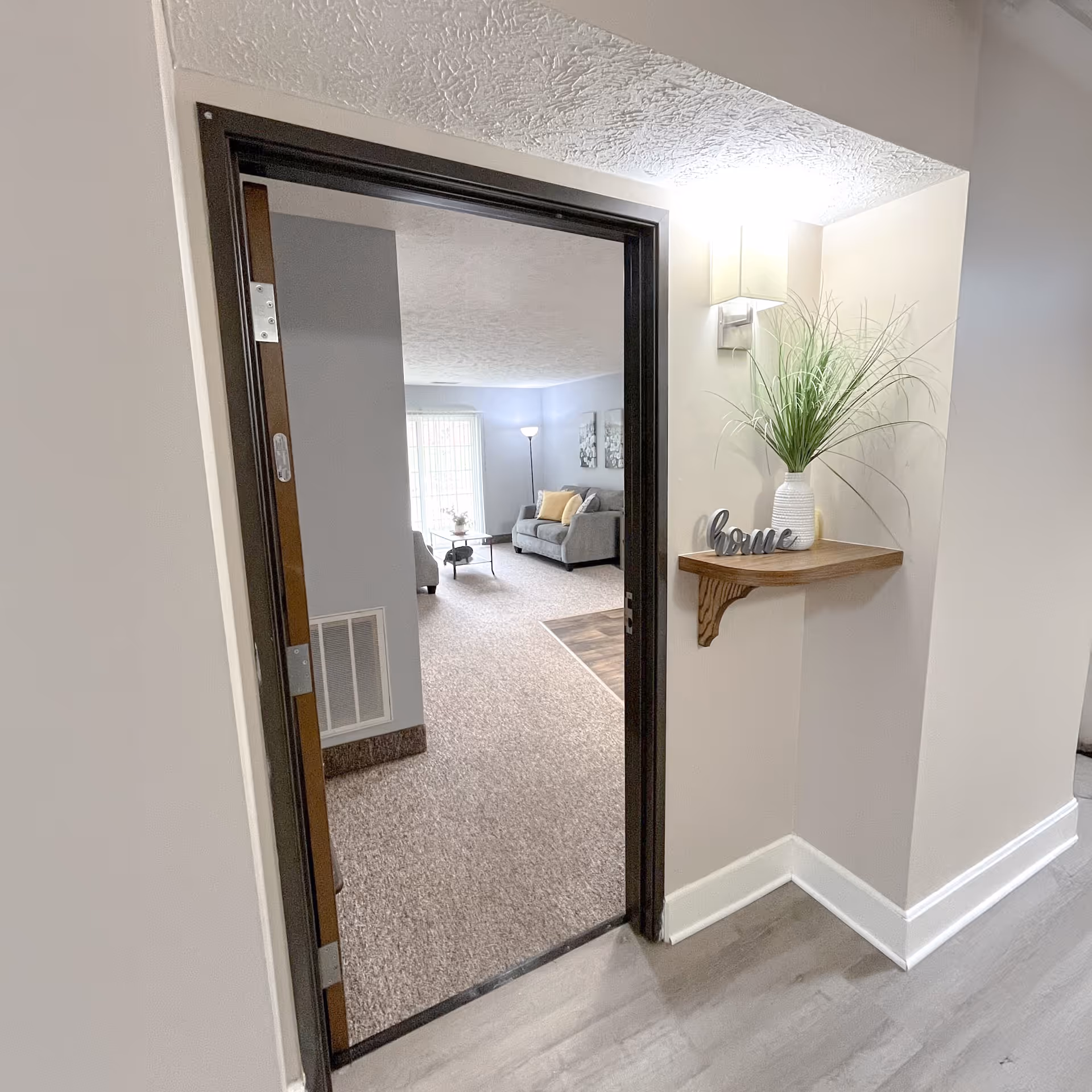 View through an open doorway into a living room with carpeted floor, a gray sofa with yellow and gray cushions, a coffee table, and a floor lamp near a window with vertical blinds. On the right side of the doorway, a small wooden corner shelf holds a white vase with green decorative grass and a decorative word sign that says 'home'. The walls are painted light beige and the floor outside the doorway is wood laminate.