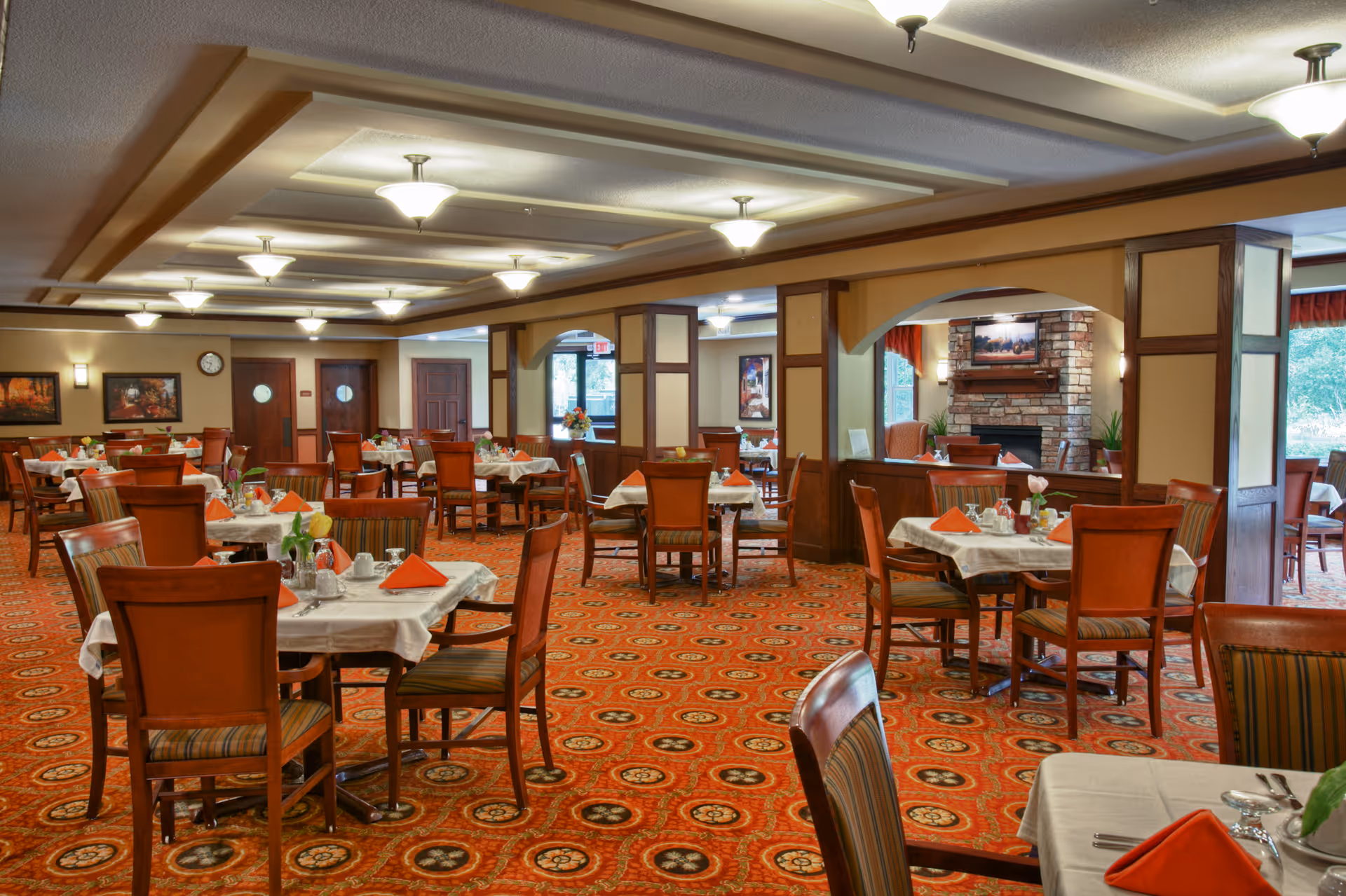 Wide dining room with multiple set tables and wooden chairs on a patterned red carpet and a fireplace in the background.