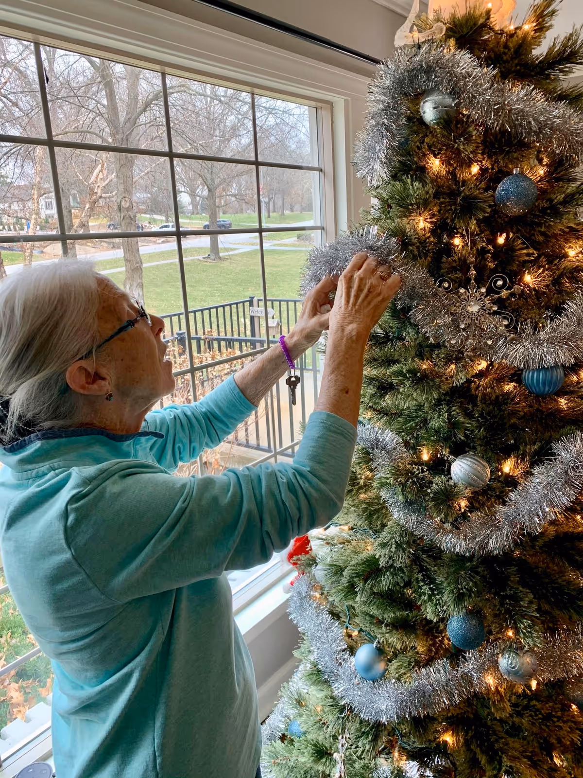 An elderly woman with gray hair tied back is decorating a Christmas tree with silver tinsel and blue ornaments near a large window showing a view of trees and a grassy area outside.