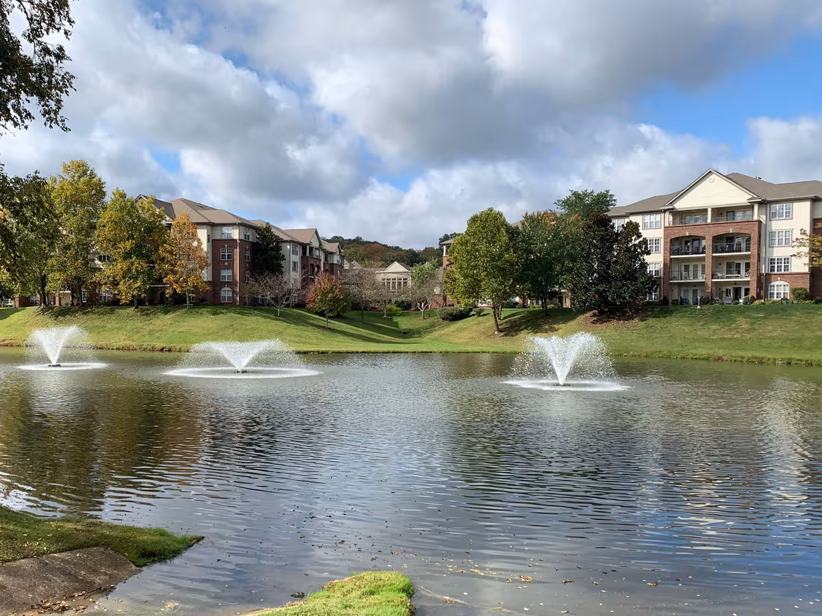 View of a pond with three water fountains in front of a senior living facility building surrounded by green grass and trees under a partly cloudy sky.
