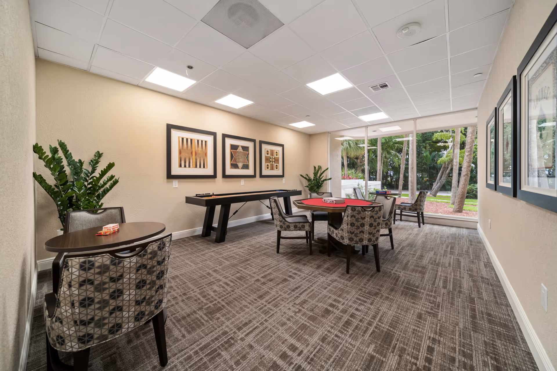 A well-lit game room with patterned carpet and beige walls, featuring a shuffleboard table against the wall, a round card table with four patterned chairs, and a small oval table with two chairs. Large windows provide a view of green trees and outdoor landscaping. Framed artwork and potted plants decorate the room.
