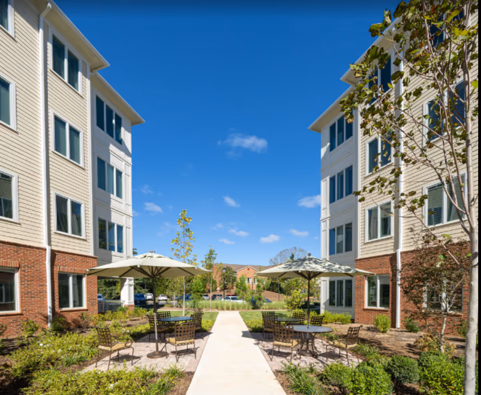 Outdoor courtyard area between two multi-story residential buildings with beige siding and brick accents. The courtyard features a paved walkway, patio tables with umbrellas, and metal chairs surrounded by landscaped greenery and small trees under a clear blue sky.