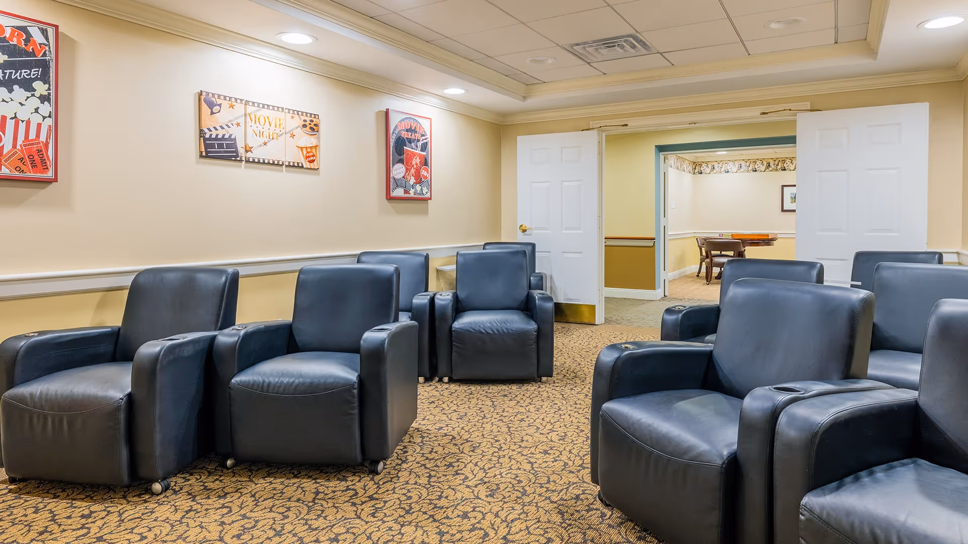 A cozy room with several black leather recliner chairs arranged in rows facing forward. The walls are decorated with movie-themed artwork, and there is a patterned carpet on the floor. Double white doors at the back of the room open into another room with a table and chairs.