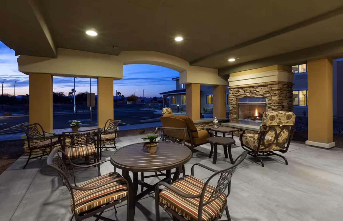 Covered outdoor seating area at dusk with multiple round tables and cushioned chairs arranged around a stone fireplace with a fire burning. The area is supported by large columns and overlooks a parking lot and distant trees under a blue evening sky.