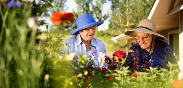 Two elderly women wearing sun hats are smiling and enjoying gardening together in a lush garden with colorful flowers and greenery, outside a residential building.