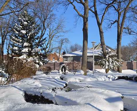 Snow blankets the front yard and trees around a residential building, partially fenced.