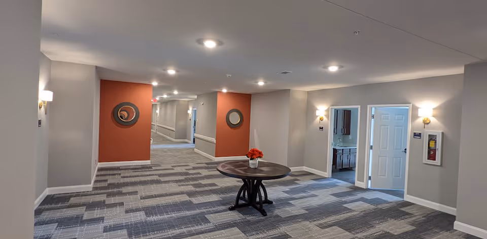 A wide hallway in an assisted living facility with patterned carpet flooring, light gray walls, and two orange accent walls each featuring a round mirror. A round wooden table with a vase of red flowers is centered in the hallway. There are two open doorways on the right side leading to rooms with cabinetry and a fire extinguisher mounted on the wall. Ceiling lights and wall sconces illuminate the space.