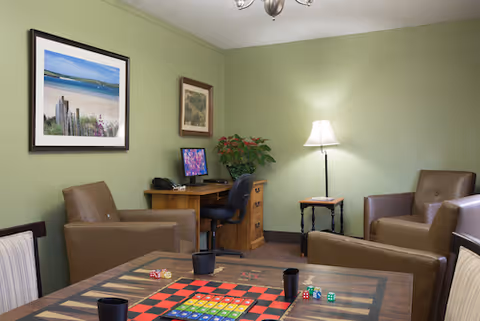 Cozy common room with a board game table in the foreground, several armchairs, a desk with a computer, framed artwork on green walls, and a floor lamp.