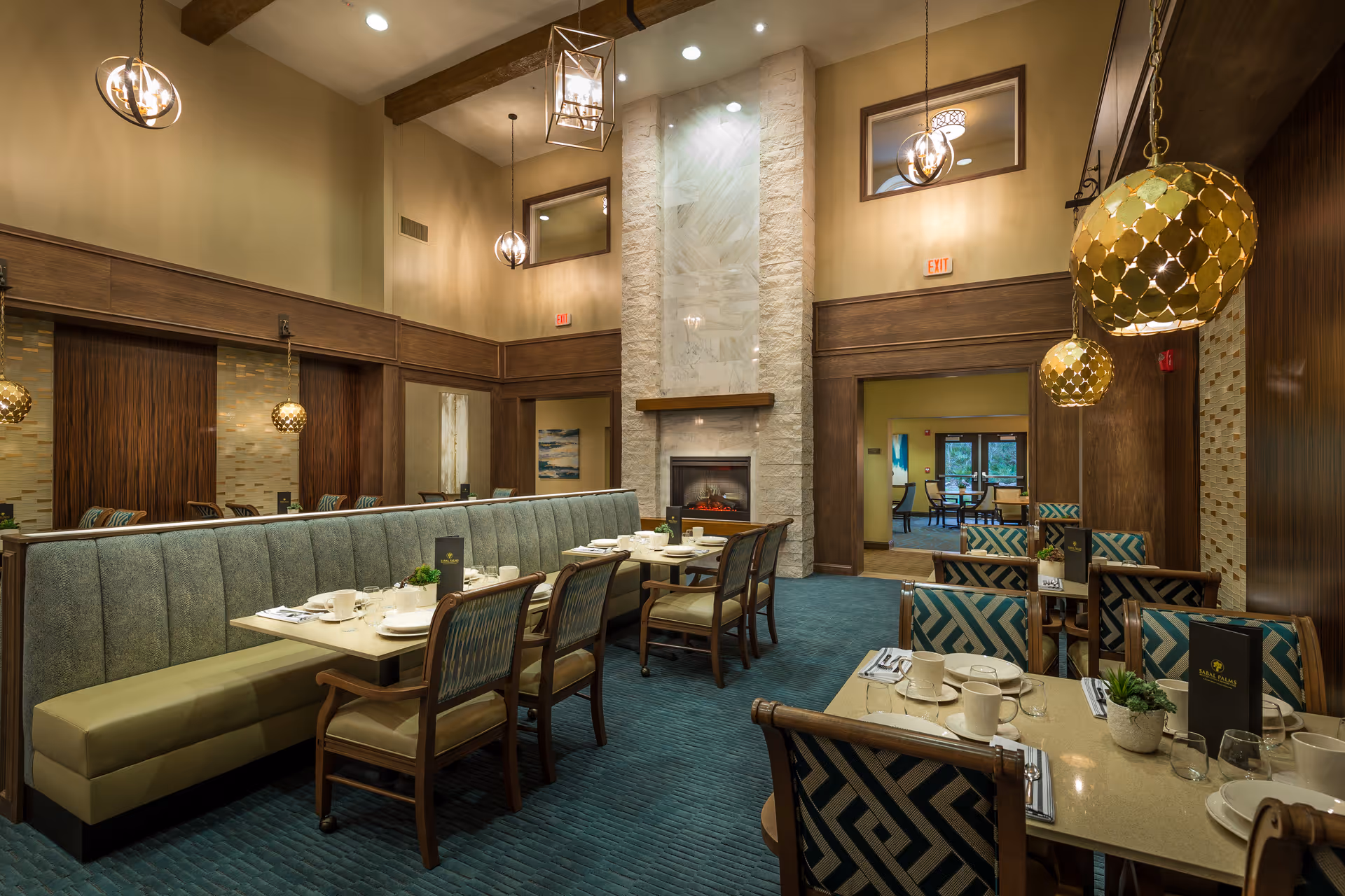 Spacious, elegantly furnished dining room with tables and chairs, a long upholstered banquette, pendant lighting, and a tall stone fireplace.