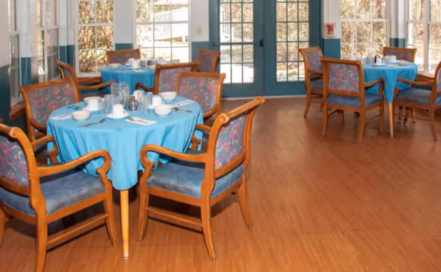 A dining room with round tables covered in blue tablecloths, each set with white cups, saucers, and utensils. Wooden chairs with floral upholstery surround the tables. Large windows and glass doors allow natural light to fill the room.