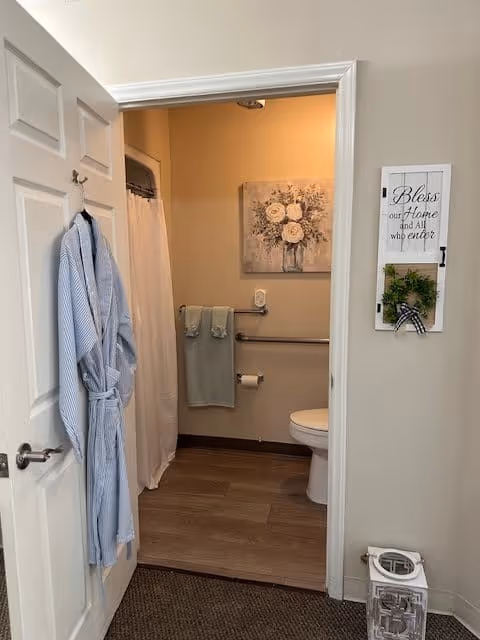 View into a bathroom from an open door with a light blue bathrobe hanging on the door. Inside the bathroom, there is a white shower curtain, a toilet, two light blue towels hanging on a grab bar, a toilet paper holder, and a floral painting on the wall. On the wall outside the bathroom, there is a decorative sign that reads 'Bless our Home and All who enter' with a small green wreath below it. A white lantern-style decorative item is on the floor near the wall.