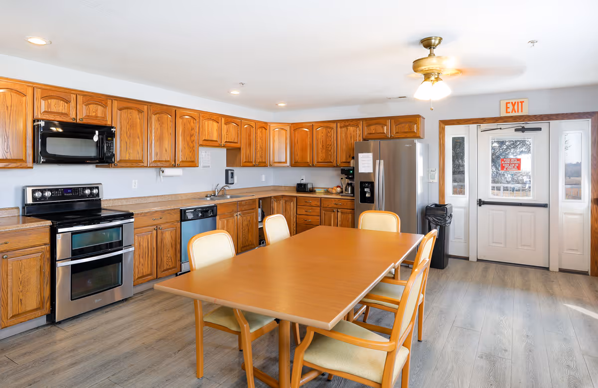A spacious kitchen area with wooden cabinets, stainless steel appliances including a double oven, microwave, dishwasher, and refrigerator. There is a wooden table with four chairs in the center of the room. The floor is light-colored wood, and there is a ceiling fan with lights. A door with an exit sign above it is visible in the background.