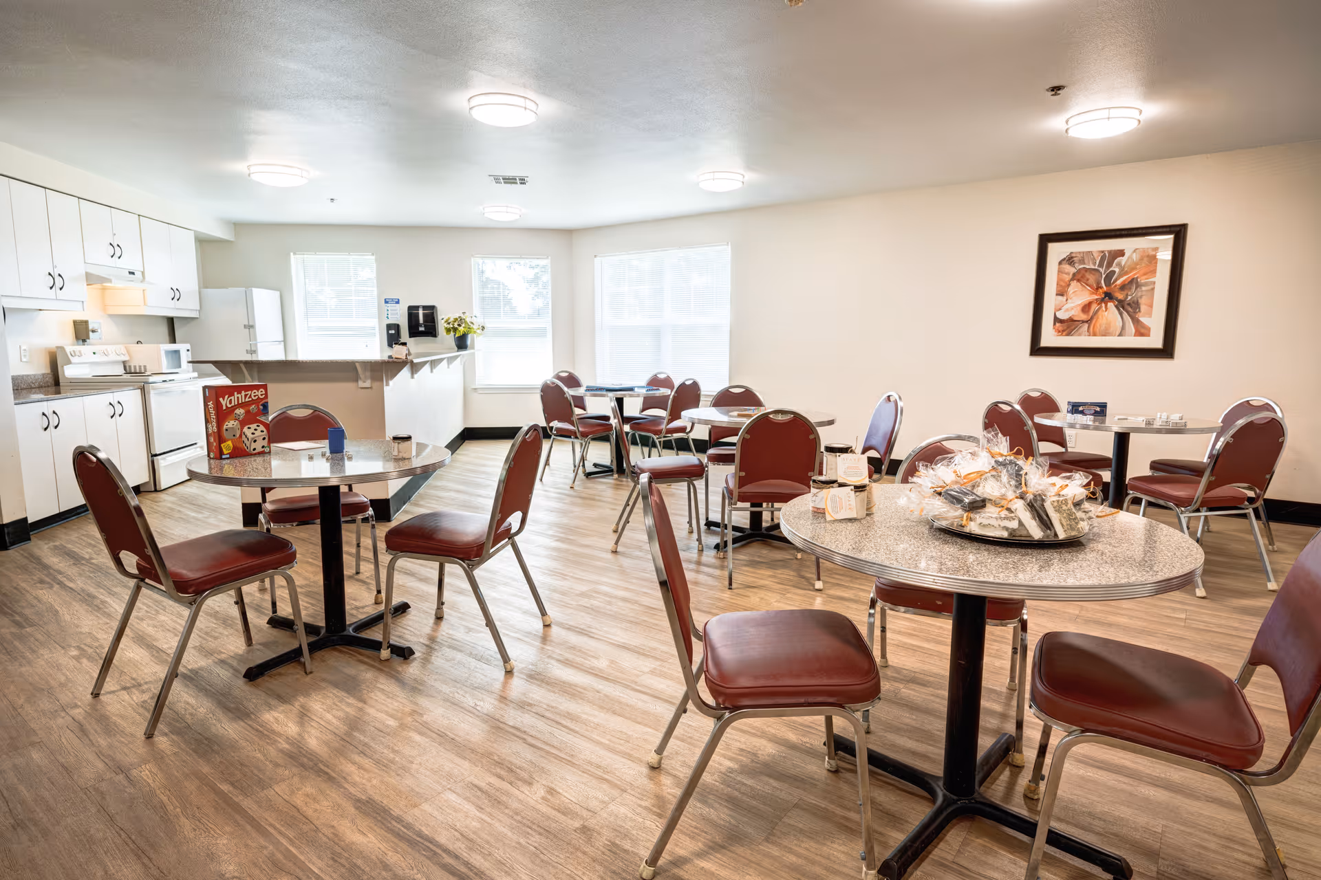 A bright communal dining/activity room with round tables and red chairs, a kitchenette in the background, and games and snacks on the tables.